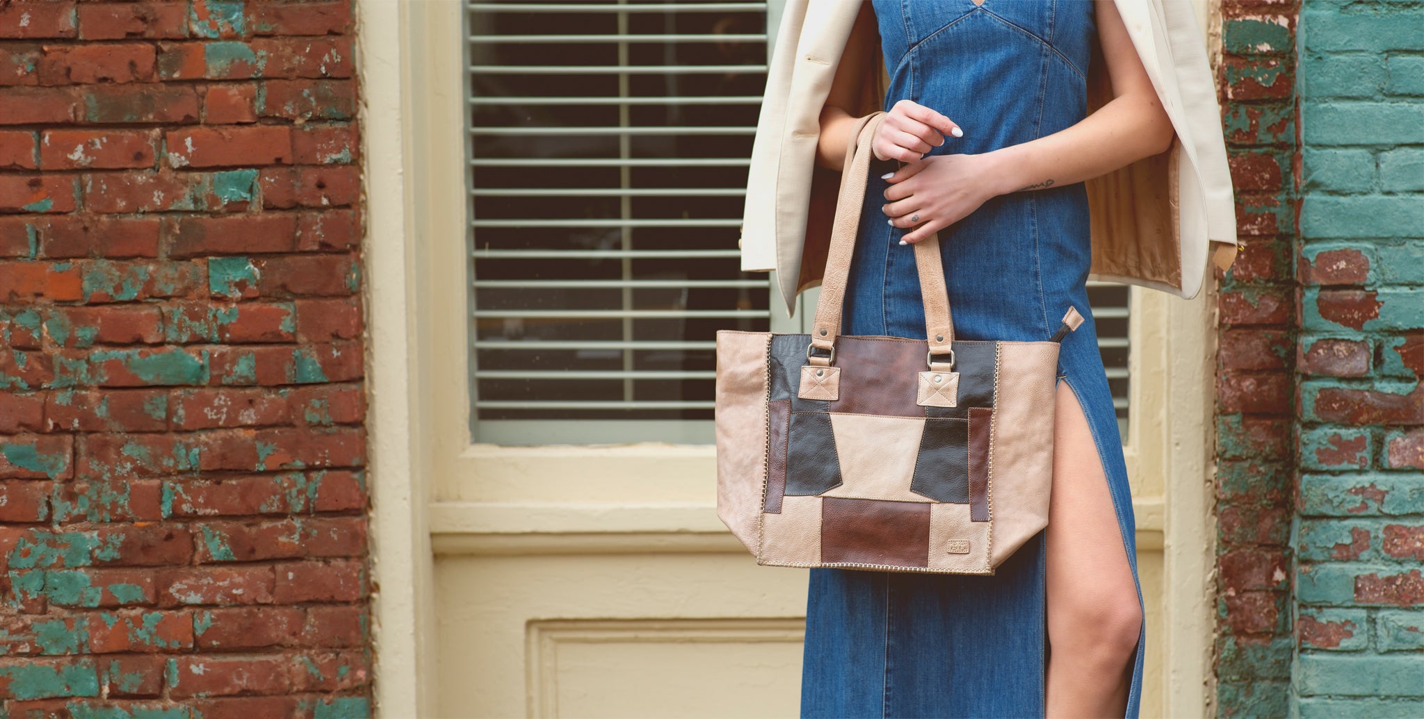 Woman in a denim dress holding a patchwork tote bag, standing against a brick wall background.