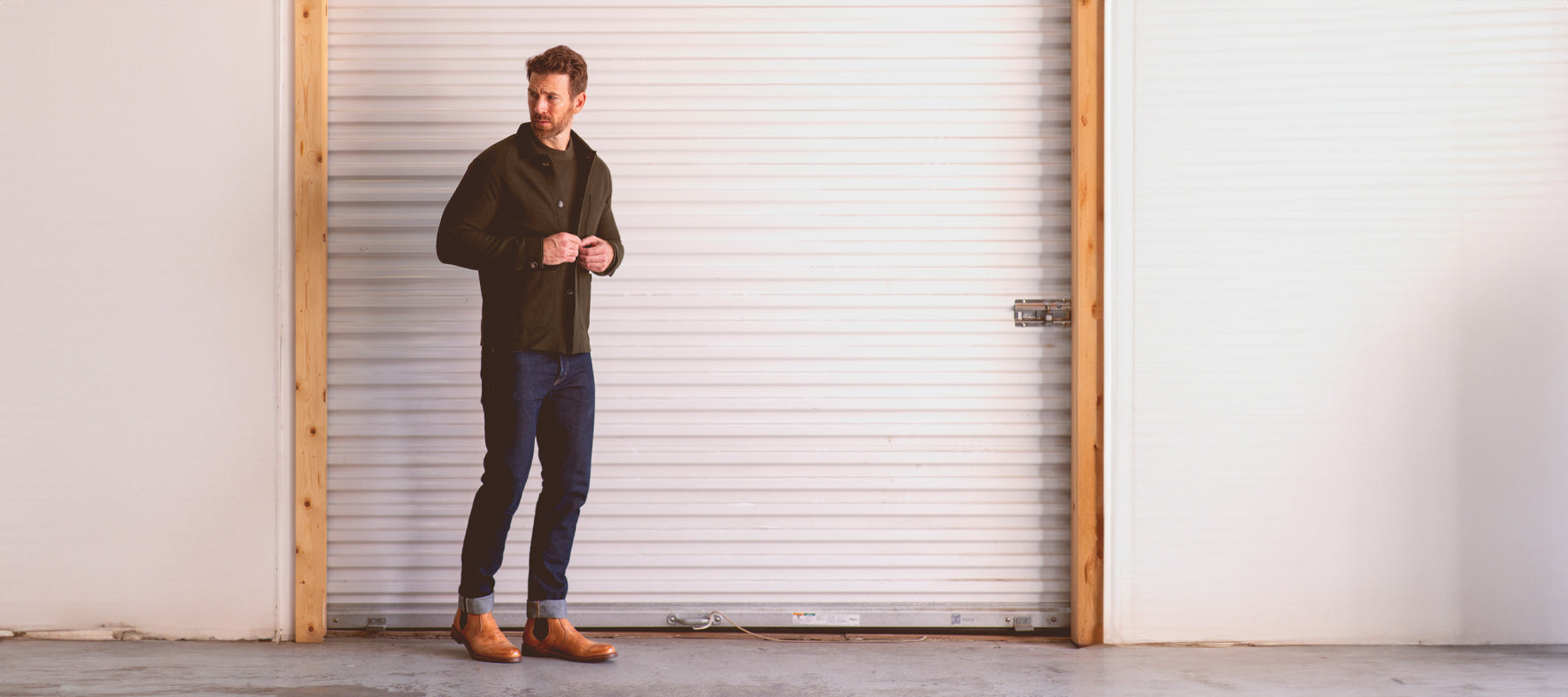 A brunette man standing in a green long sleeve shirt and cuffed jeans  and tan boots in front of a white garage door.