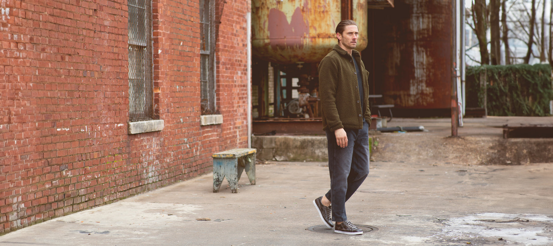 A man with brown hair slicked back wearing a green jacket, navy chinos and black leather sneaakers in a outside rusted warehouse and near a rustic brick wall.