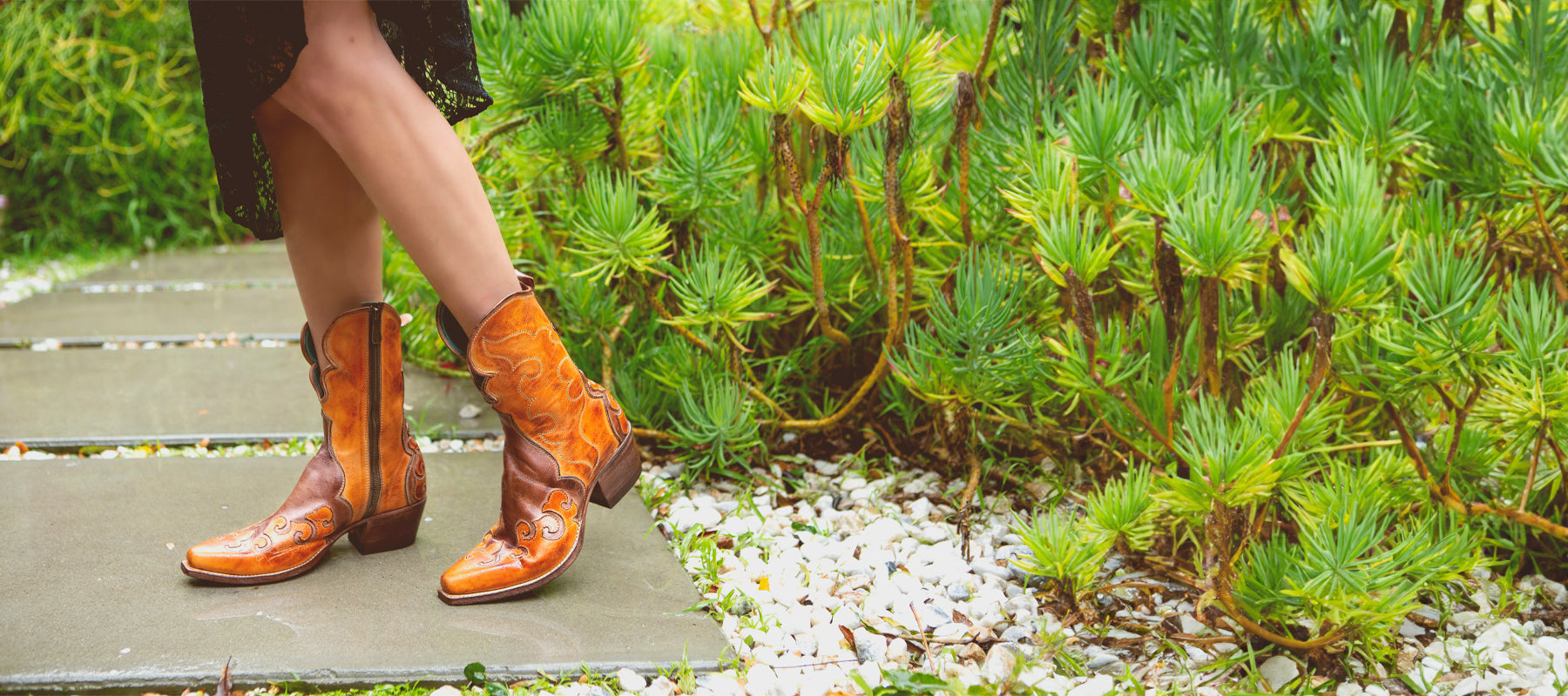 Person wearing detailed cowboy boots standing beside a garden path with lush greenery.