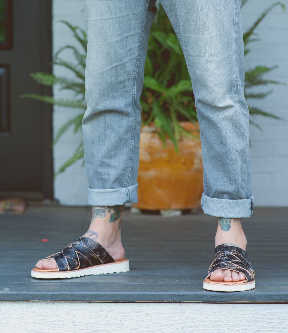 A person wearing Abraham Light jeans and Bed Stu black leather sandals stands on a white doorstep, revealing tattooed ankles.