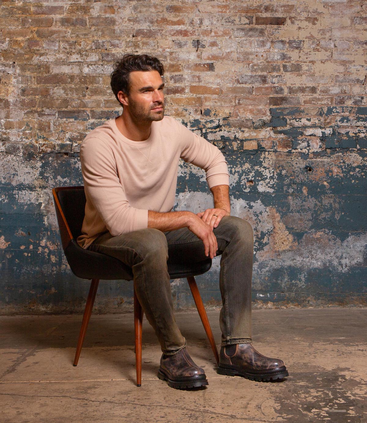 A man with dark hair and a beard sits on a chair against a brick wall, wearing a beige long-sleeve shirt, gray jeans, and Bed Stu Accelerate men’s Chelsea boots featuring a rugged lug outsole for a modern twist.