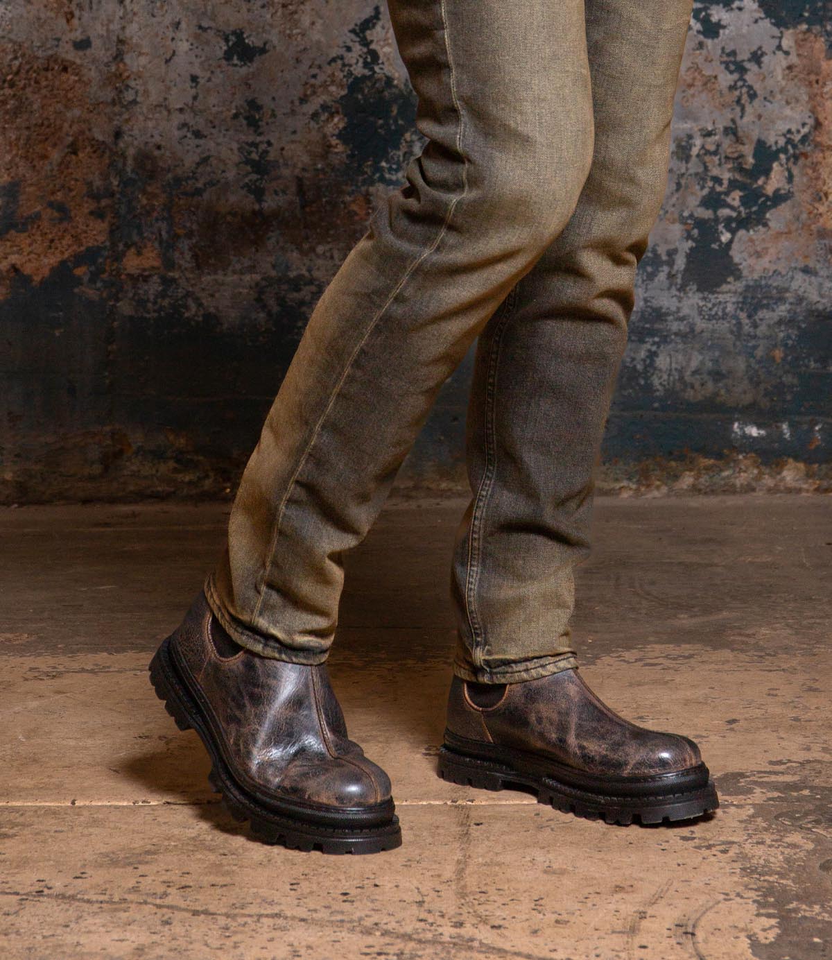 A person wearing faded jeans and Bed Stu’s Accelerate men’s Chelsea boots with a rugged lug outsole stands on concrete against a textured wall, giving the classic look a modern twist.