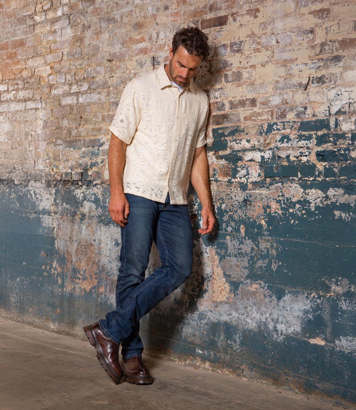 A man in a light short-sleeve shirt, jeans, and Bed Stu Accelerate men's Chelsea boots with rugged lug outsoles stands against a weathered brick wall, looking down with his hands at his sides.