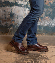 A person in blue jeans and Bed Stu’s Accelerate dark brown Chelsea boots with a rugged lug outsole stands on worn concrete against a weathered wall, giving classic style a modern edge.