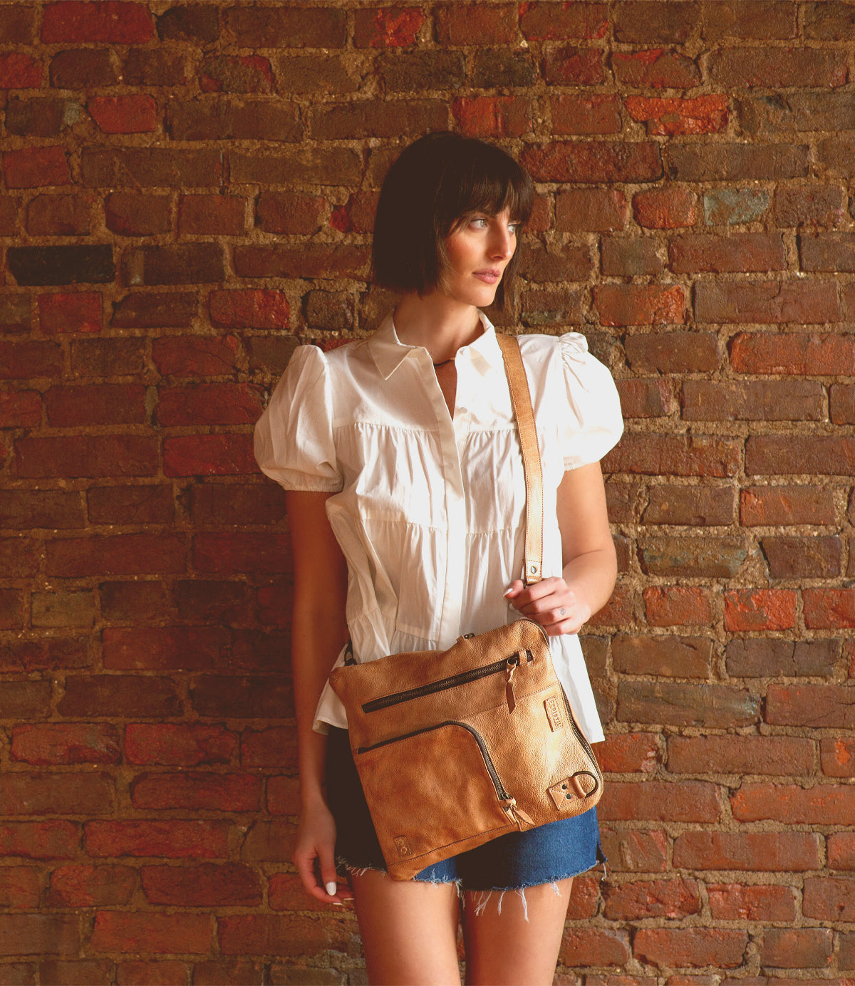 A woman is standing in front of a brick wall holding a Bed Stu Aiken convertible crossbody bag.