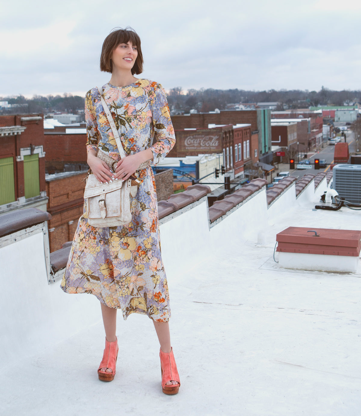 A woman in a floral dress and orange heels stands on a rooftop, holding a Bed Stu Ainhoa LTC crossbody handbag, with a small town backdrop under a cloudy sky.