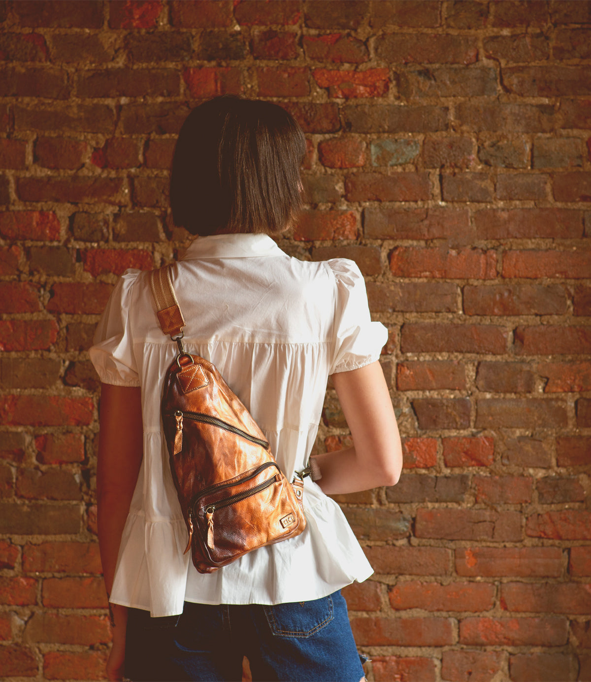 A practical woman with the versatile Bed Stu Andie leather sling bag poses in front of a brick wall.
