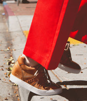 A person wearing bright red pants and the Bed Stu Andromeda tan leather high-top sneakers with a street-smart design is stepping off a curb onto the street.