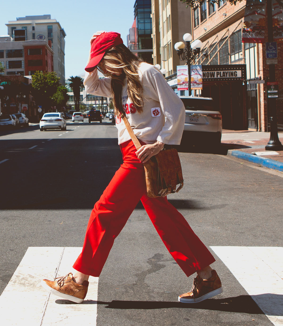 A person crossing a street in an urban area, wearing a red cap, white long-sleeve shirt, red pants, and brown shoes with hidden heels. Slung over their shoulder is the Andromeda satchel by Bed Stu, completing this street-smart design.