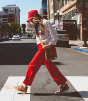 A person crossing a street in an urban area, wearing a red cap, white long-sleeve shirt, red pants, and brown shoes with hidden heels. Slung over their shoulder is the Andromeda satchel by Bed Stu, completing this street-smart design.