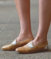 A person wearing light brown, two-tone leather Ballet shoes by Bed Stu with a white strap stands on a speckled surface, exuding both comfort and flexibility.