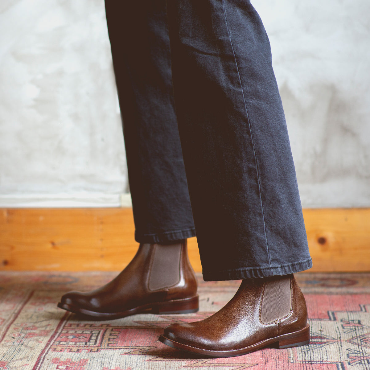 A person standing on a Barfly rug wearing brown Bed Stu Chelsea boots made of Italian leather.