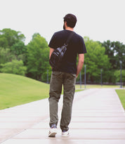 A man with a beard walks along a paved path in a park, wearing a black shirt, khaki pants, white sneakers, and carrying the Bed Stu Beau leather sling backpack with a durable strap. Green trees and grass line the background.