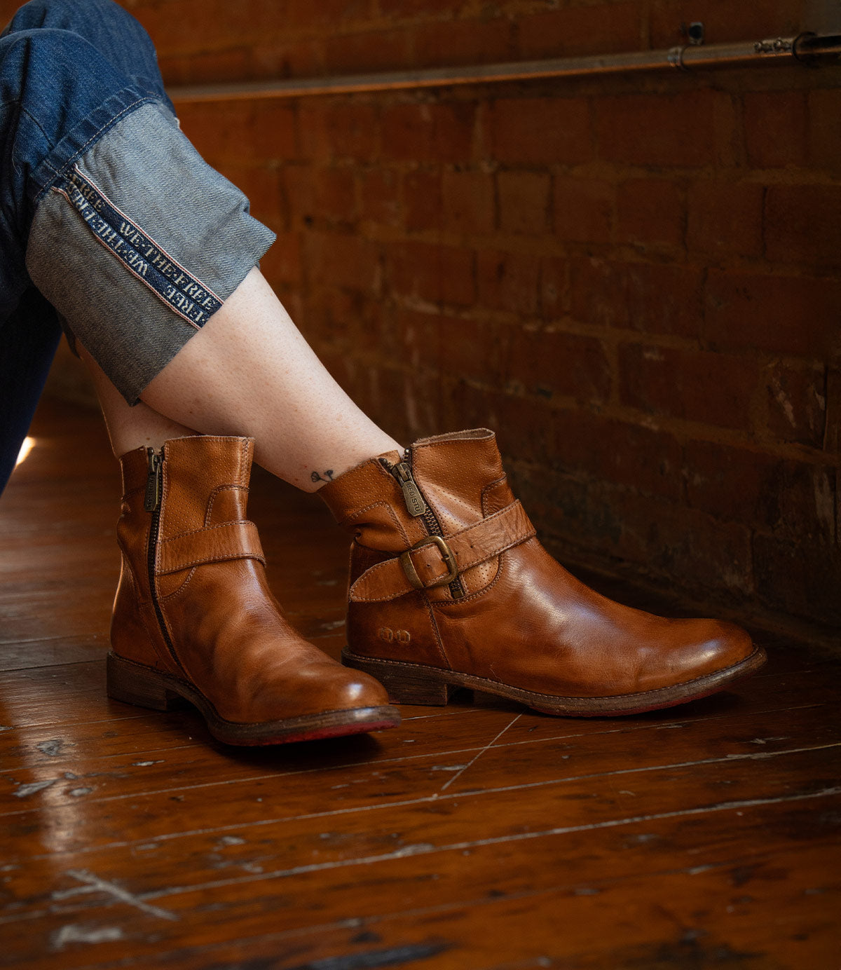 A person in rolled-up jeans and Bed Stu's Becca brown leather short engineer boots with dual zippers sits on a wooden floor against a brick wall.