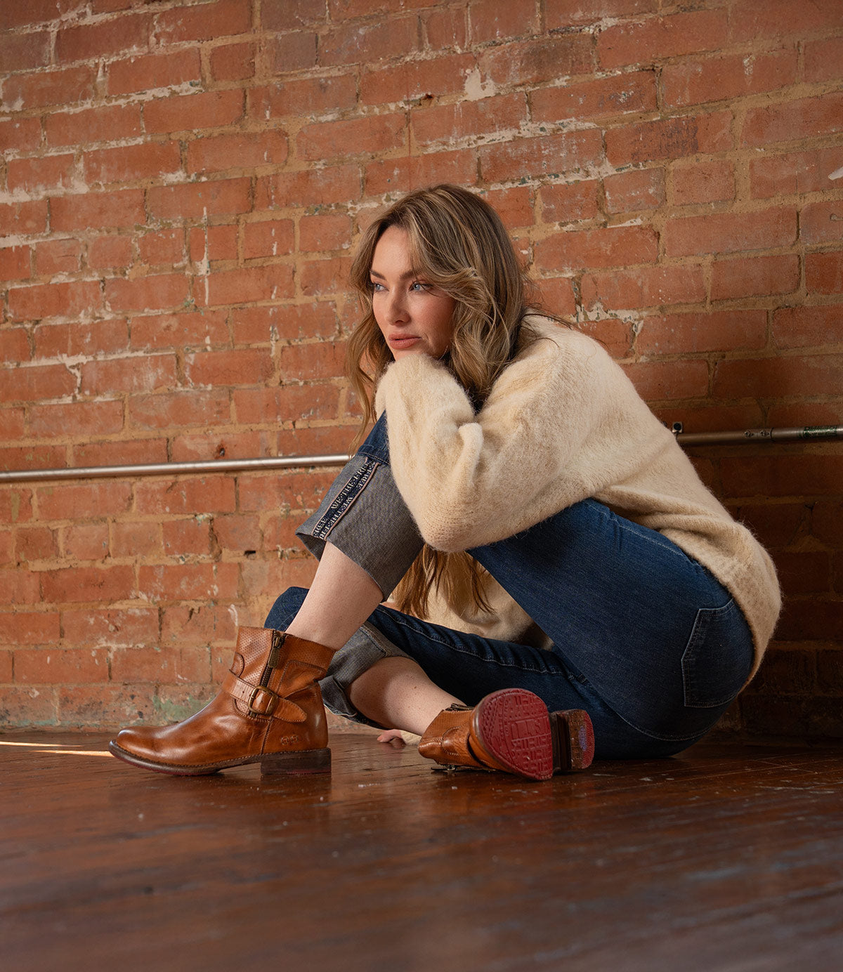 A woman wearing Bed Stu's Becca short engineer boots with dual zippers and an ankle buckle sits on a wooden floor against a brick wall, resting her arms on her knee and looking to the side.