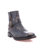 The Bed Stu Becca is a black leather short engineer boot featuring dual zippers, an ankle buckle strap, a worn finish, and a low heel, displayed against a white background.