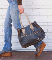 A person walking with a large, Bruna handbag by Bed Stu, featuring dark blue color, decorative stitching, and padded shoulder straps. They are wearing blue jeans and brown wedge-heeled shoes, against a white brick wall background.