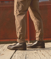 Someone wearing beige pants and Bed Stu's Barton leather high-top sneakers stands on a wooden surface with a red fence in the background.