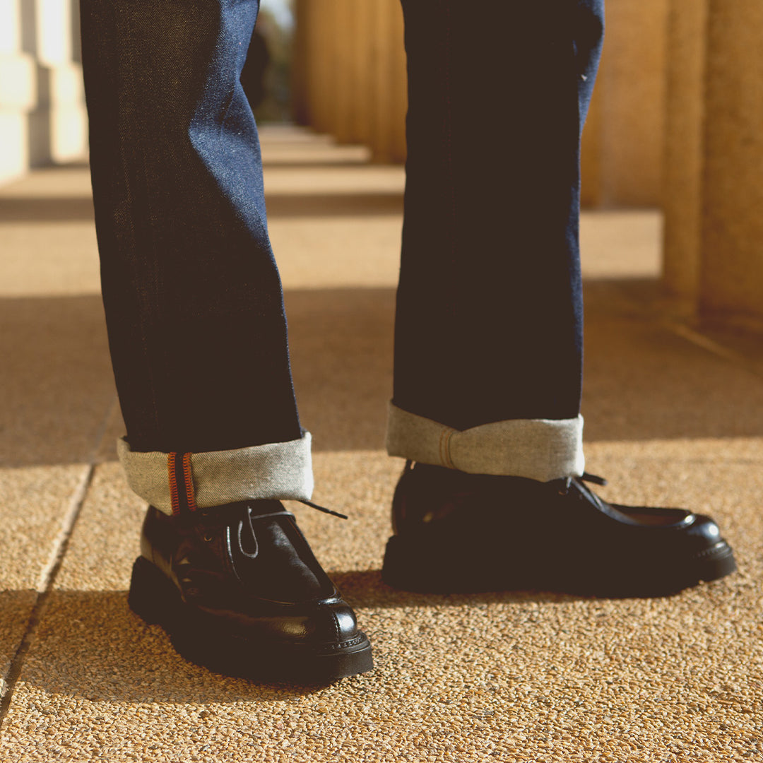 A person stands on textured ground wearing cuffed jeans and sleek Casotto moc-toe lace-up boots from Bed Stu, expertly crafted from Italian vegetable-tanned leather.