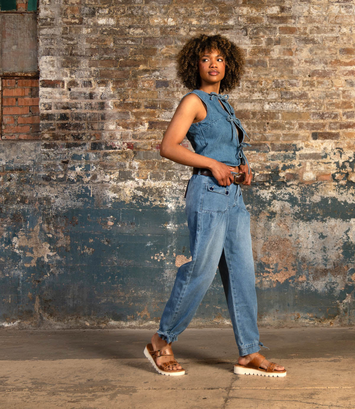 A woman wearing the Bed Stu Chow sleeveless denim jumpsuit and cushioned sandals stands in front of a weathered brick wall, looking to the side.