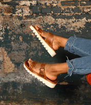 A person wearing Bed Stu's Chow brown leather slide sandals with a chunky rubber sole and blue jeans stands in front of a textured, weathered wall.