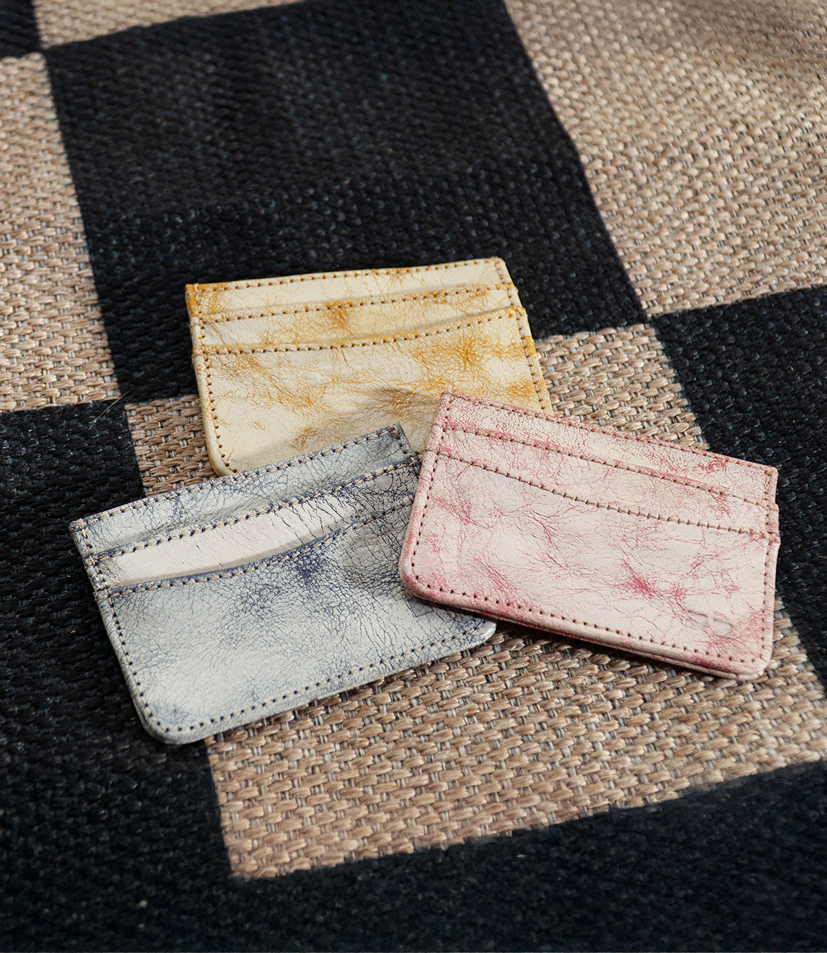 Three Chuck leather card holders by Bed Stu in yellow, pink, and gray rest elegantly on a woven checkered fabric background—ideal for organizing your credit cards or business cards with style.