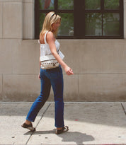 A woman with blonde hair walks on a sidewalk in front of a building, sporting a white top, blue jeans, brown sandals, and a versatile Bed Stu Cleo crossbody bag with a zipper top closure. She is looking down and slightly smiling.