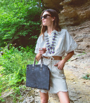 A woman wearing a white shirt and sunglasses holding a Bed Stu Commuter black tote bag.