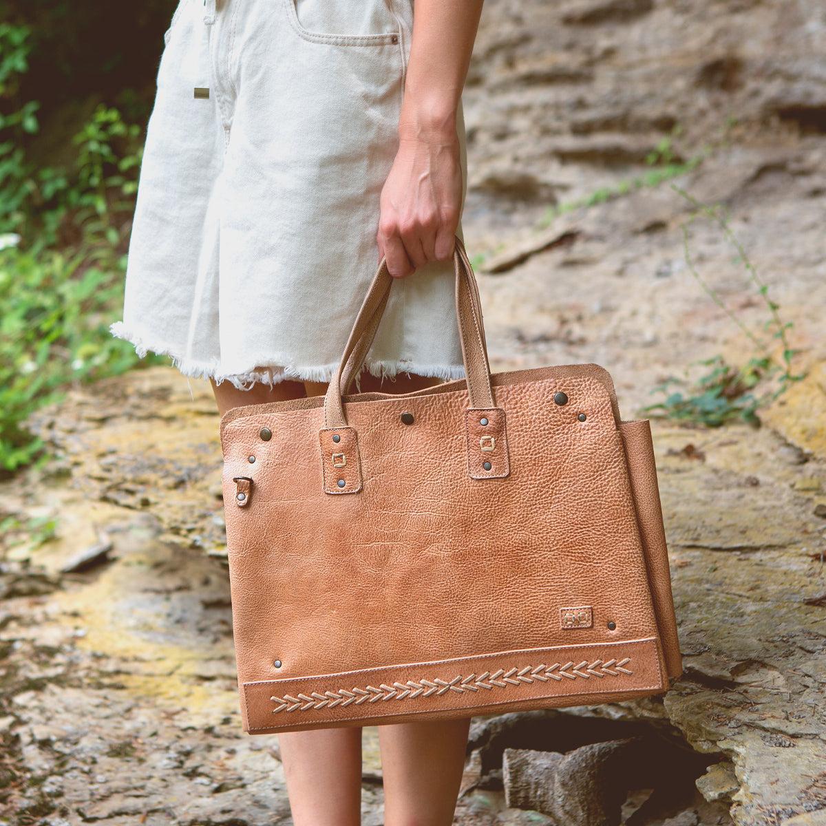A woman is holding a multifunctional Bed Stu tan leather bag perfect for commuters with laptops.