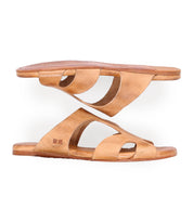 A pair of brown slip-on leather sandals with cut-out designs and textured soles, featuring a vegetable-tanned leather upper and a cushioned inner sole, stacked one on top of the other, Bed Stu Configure.