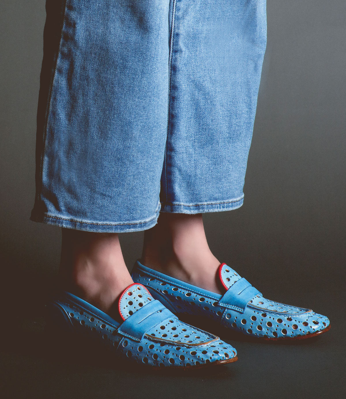 Person wearing light blue jeans and light blue Cornflower loafers by Bed Stu with a playful cutout pattern and red accents, standing against a dark background. These handcrafted in Italy shoes exude style and elegance.