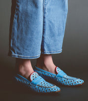 Person wearing light blue jeans and light blue Cornflower loafers by Bed Stu with a playful cutout pattern and red accents, standing against a dark background. These handcrafted in Italy shoes exude style and elegance.