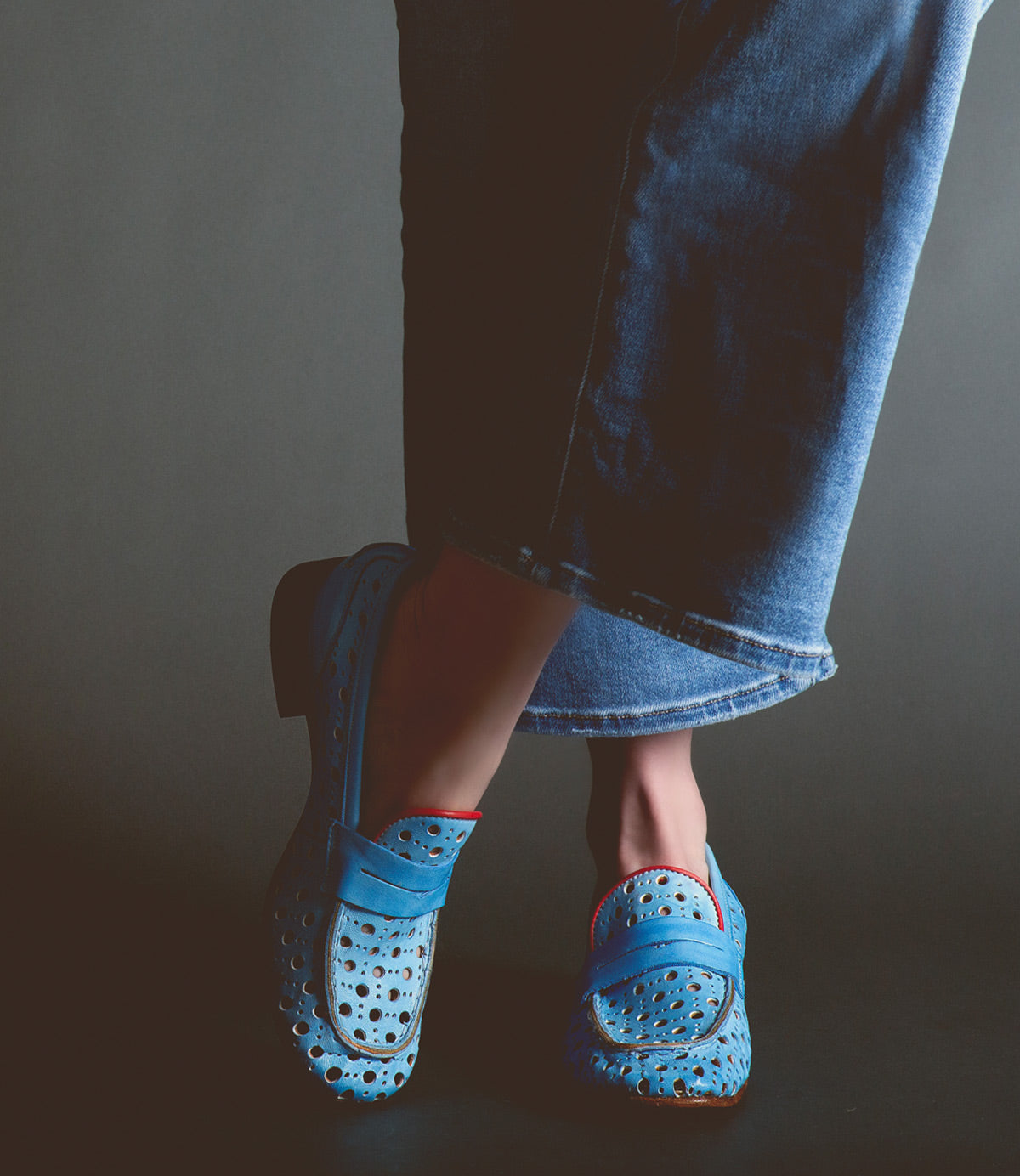 A person wearing Bed Stu Cornflower penny loafers with a circular cutout pattern and denim jeans against a dark background.