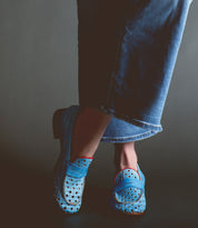 A person wearing Bed Stu Cornflower penny loafers with a circular cutout pattern and denim jeans against a dark background.
