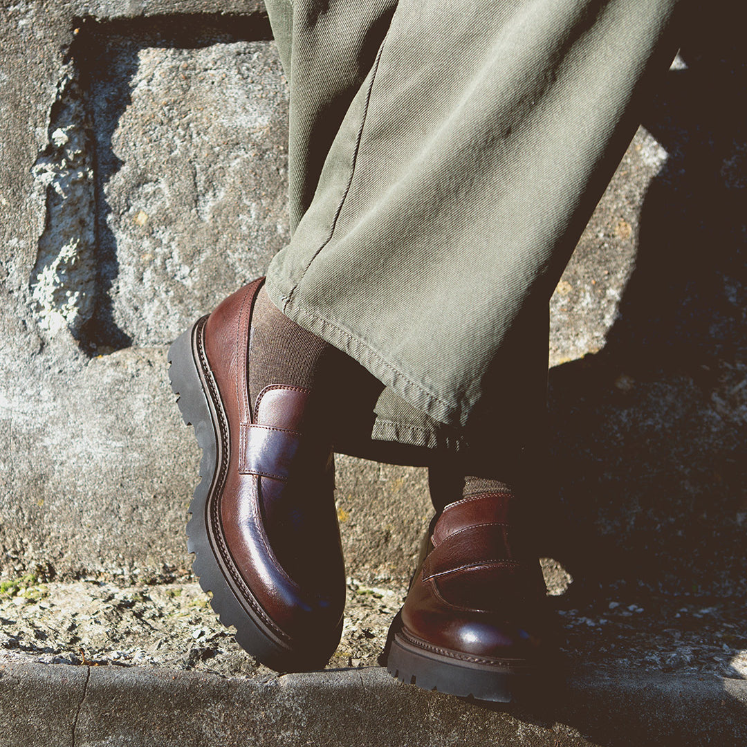 A person wearing Bed Stu's Costruito loafers with a chunky Livetech outsole and olive green pants stands against a stone wall.
