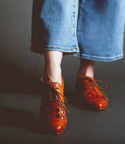 Person wearing wide-leg blue jeans and unique women's footwear in the form of Decoy by Bed Stu, brown leather oxford shoes with cut-out details, standing on a dark surface.