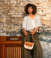 A person with curly hair stands against a brick wall, wearing a white blouse, olive pants, and holding the Bed Stu Elect shoulder bag with an adjustable strap in front of a vintage wooden radio.