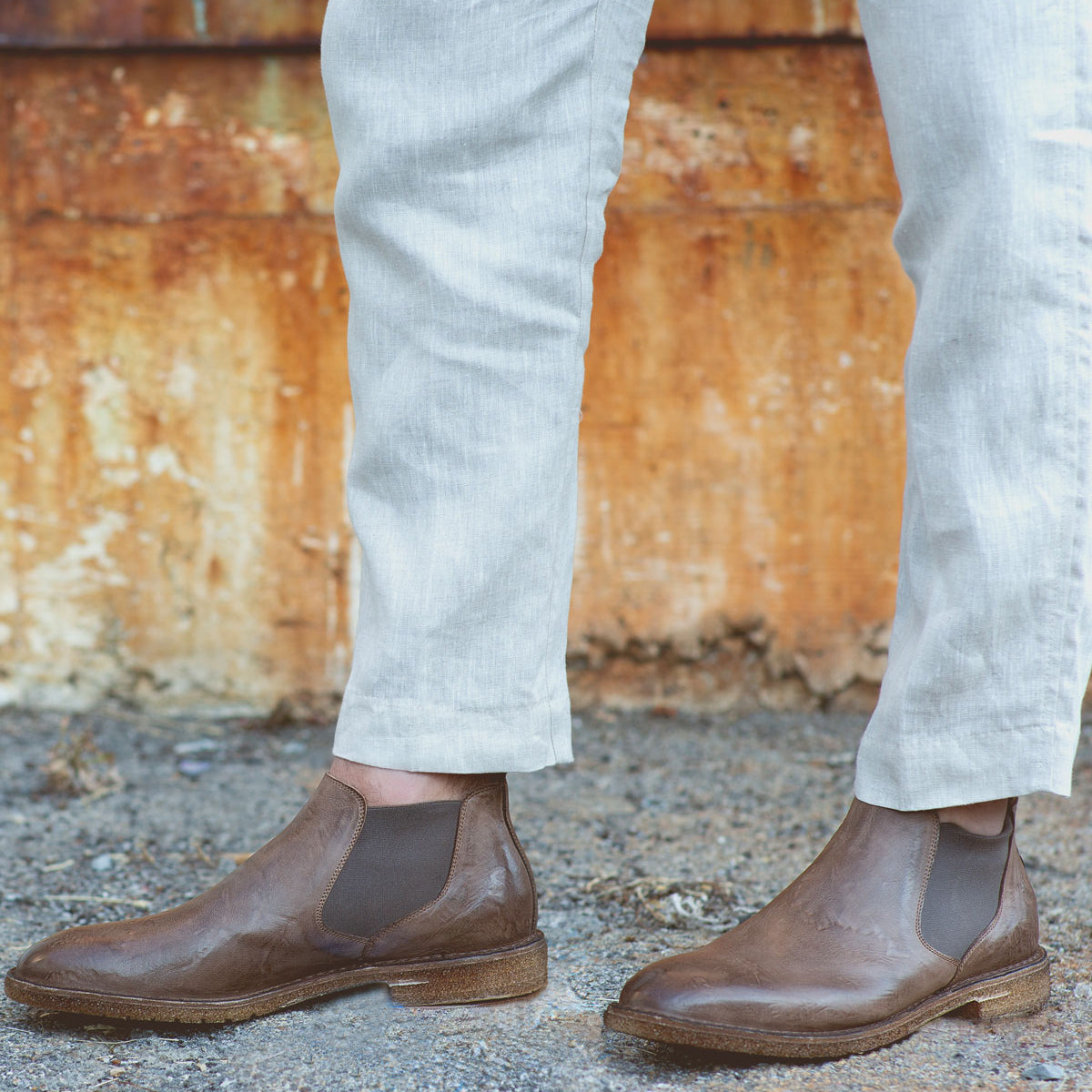 Person wearing light-colored pants and the Elude men's leather boots by Bed Stu, handcrafted in Italy, standing on gravel with a rusted metal wall in the background.