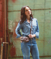A woman wearing a denim shirt and jeans standing in front of a building with a Bed Stu bag featuring leather zipper pulls.