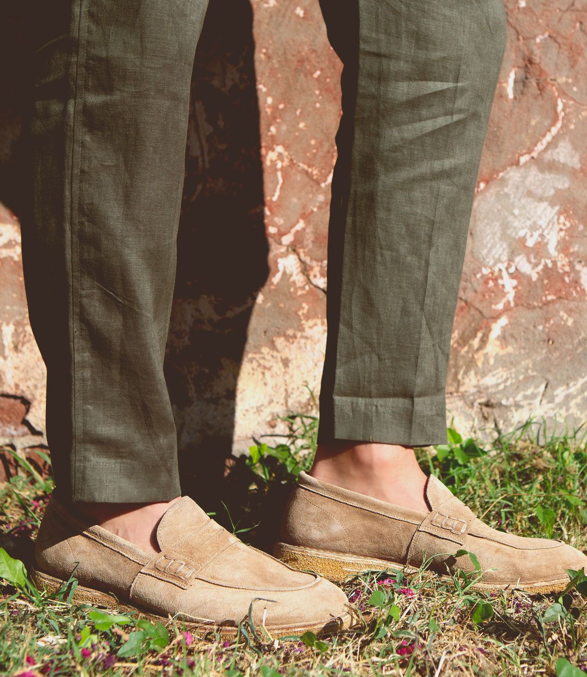 A person wearing green pants and tan suede Bed Stu Envoy loafers stands on grass with a rustic wall in the background, exuding Italian elegance.