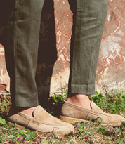 A person wearing green pants and tan suede Bed Stu Envoy loafers stands on grass with a rustic wall in the background, exuding Italian elegance.