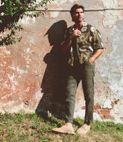 An individual stands against a weathered wall, adorned in the timeless sophistication of Bed Stu's Envoy shirt and green pants, with one hand in their pocket and the other holding a cigarette.
