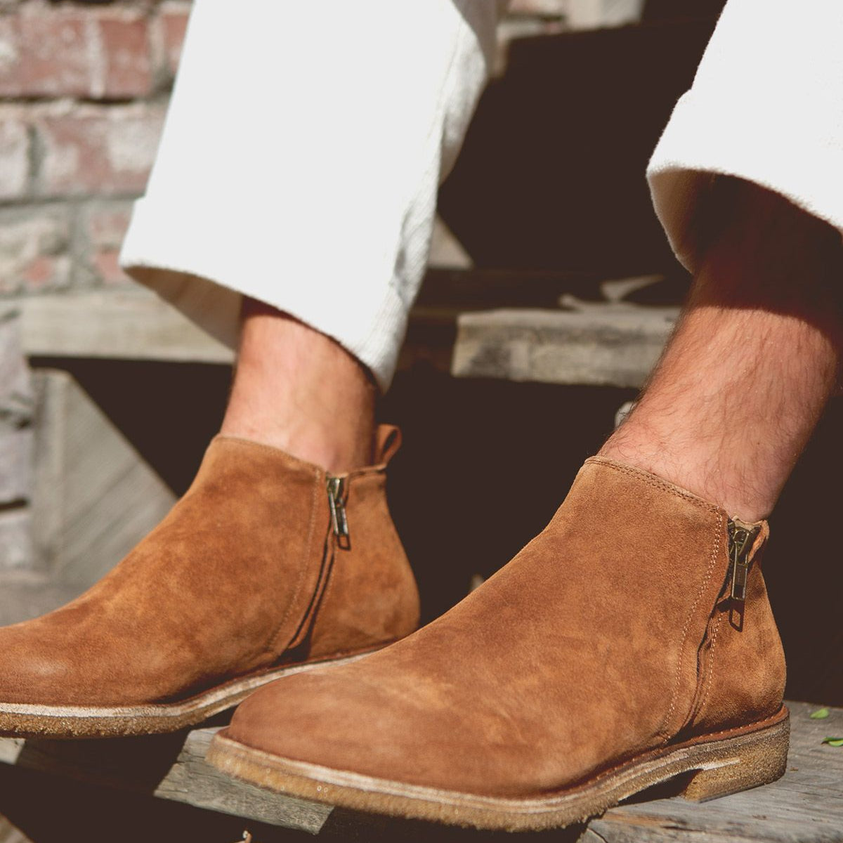 A person wearing light-colored pants and Bed Stu Evade handcrafted brown suede ankle boots with dual zippers, seated on wooden steps in an outdoor setting.