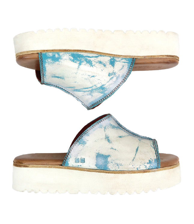 The Bed Stu Fairlee II features two white flatform slide sandals with blue tie-dye patterns, open toes, and a leather-lined footbed for added comfort, shown from the side and above.