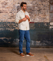 A man stands indoors by a weathered brick wall, wearing a light button-up shirt, blue jeans, and Bed Stu Fairman low-top leather sneakers featuring a comfortable insole.
