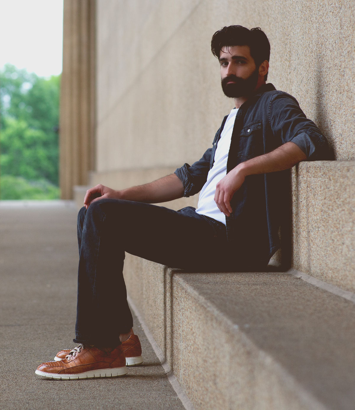 A man with a beard, wearing a denim jacket featuring unique topstitching, a white shirt, and brown **Fairman** shoes by **Bed Stu**, sits on a stepped ledge against a concrete wall. Greenery is visible in the background.