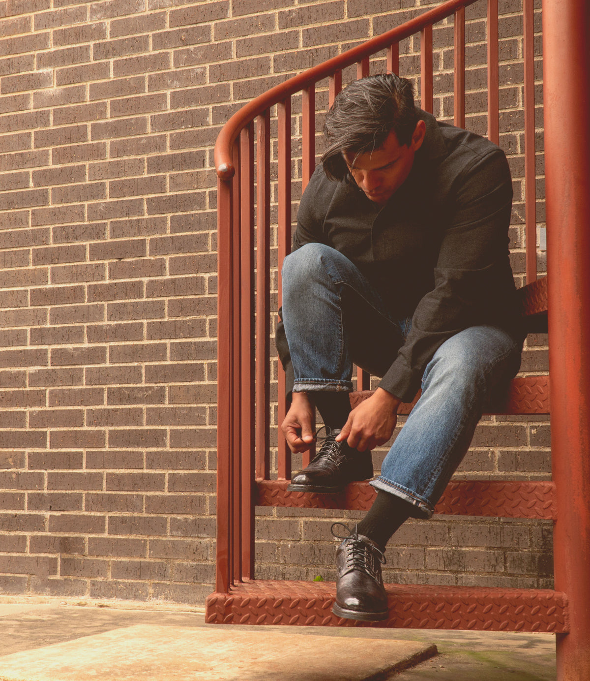 A person crouches on metal stairs, lacing up the sleek Barton men's sneaker by Bed Stu, featuring a breathable insole. They're in a black shirt and blue jeans, framed by a rustic brick wall.