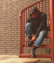 A person crouches on metal stairs, lacing up the sleek Barton men's sneaker by Bed Stu, featuring a breathable insole. They're in a black shirt and blue jeans, framed by a rustic brick wall.