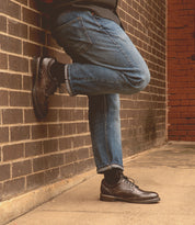 A person in jeans and Bed Stu Barton leather high-top sneakers leans against a brick wall, one leg crossed over the other.